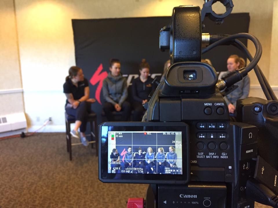 A video camera recording a panel of athletes seated in front of a sponsor backdrop